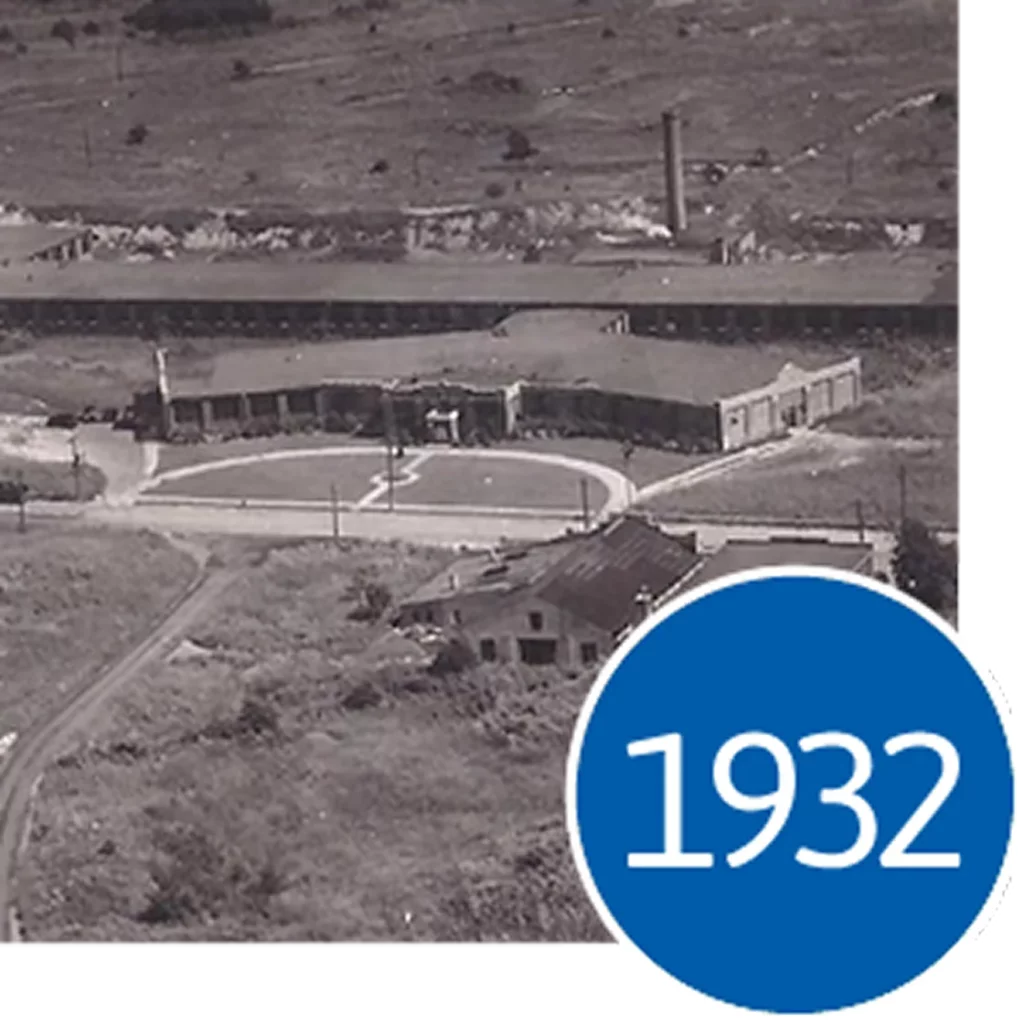 Aerial black and white photo of a 1932 industrial building complex with a circular driveway and blue text overlay.
