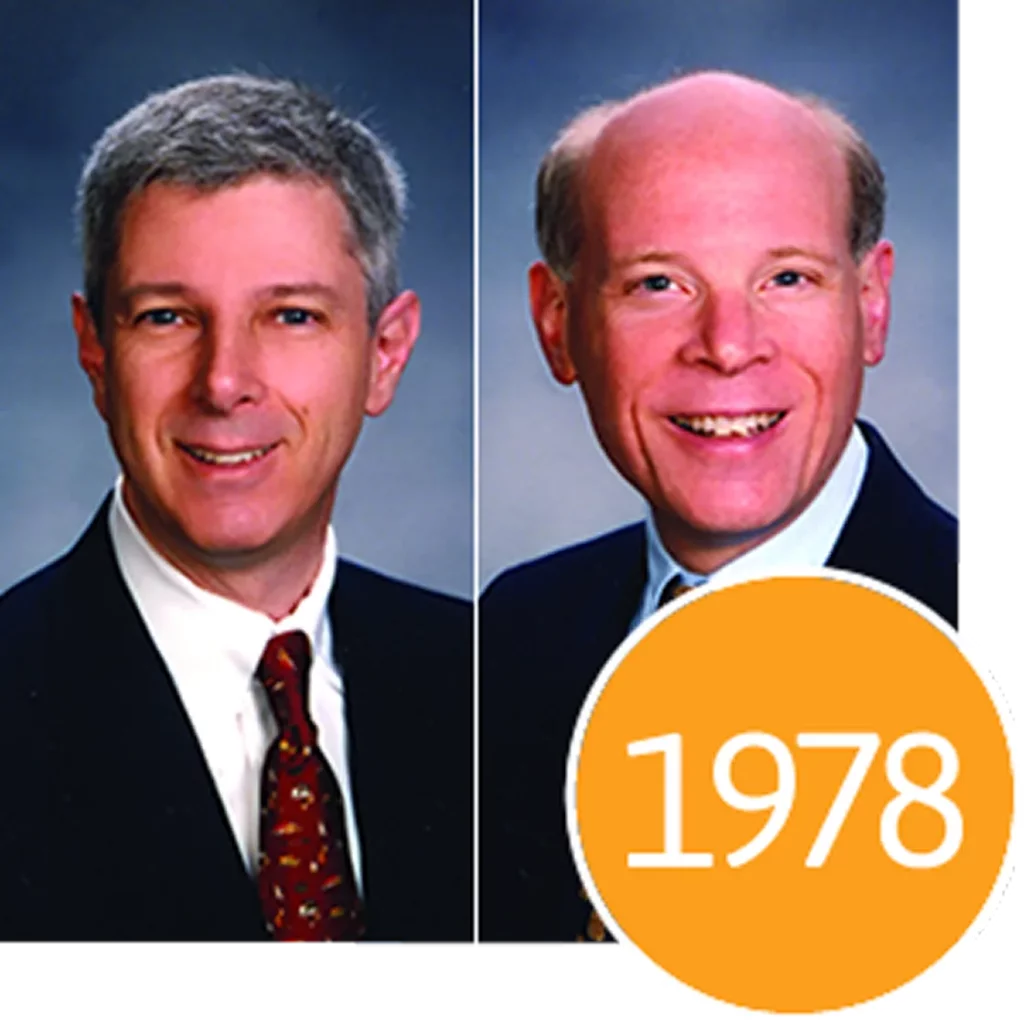 Two side-by-side portraits of smiling men in suits with a large orange circle containing the year 1978.