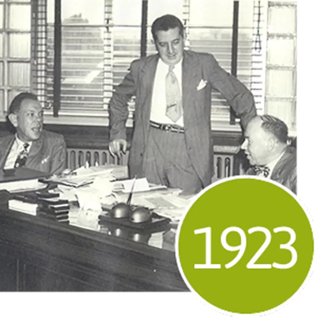 Three men in suits gather around a desk in an office with a green circle overlay reading 1923.