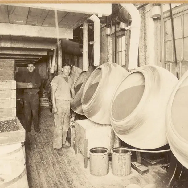 A black and white photo of two men in a factory standing beside a row of large, tilted, rotating industrial drums.