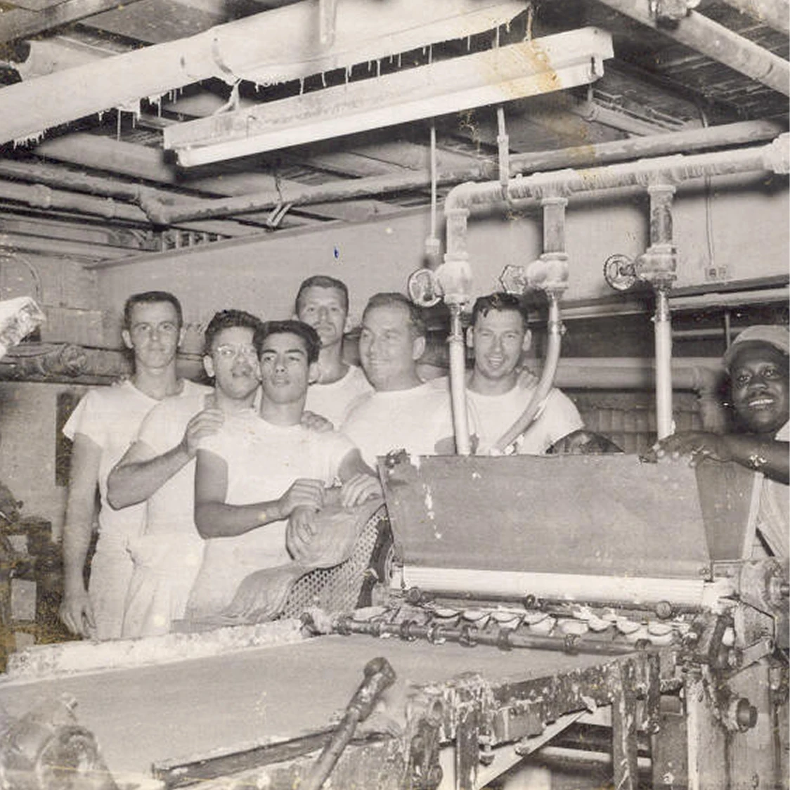 Vintage black and white photo of seven men in white t-shirts posing behind industrial factory machinery.