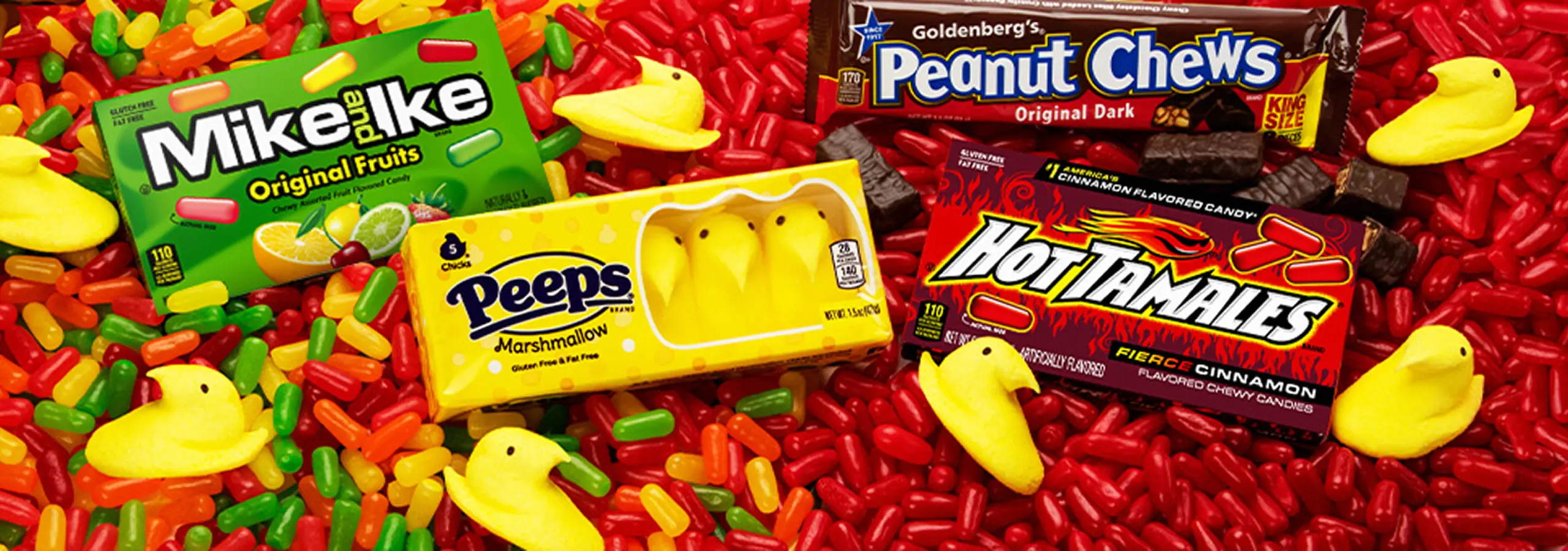 An assortment of various candies, including boxes of Mike and Ike, Peeps, Hot Tamales, and Peanut Chews, scattered over a colorful bed of loose candies.