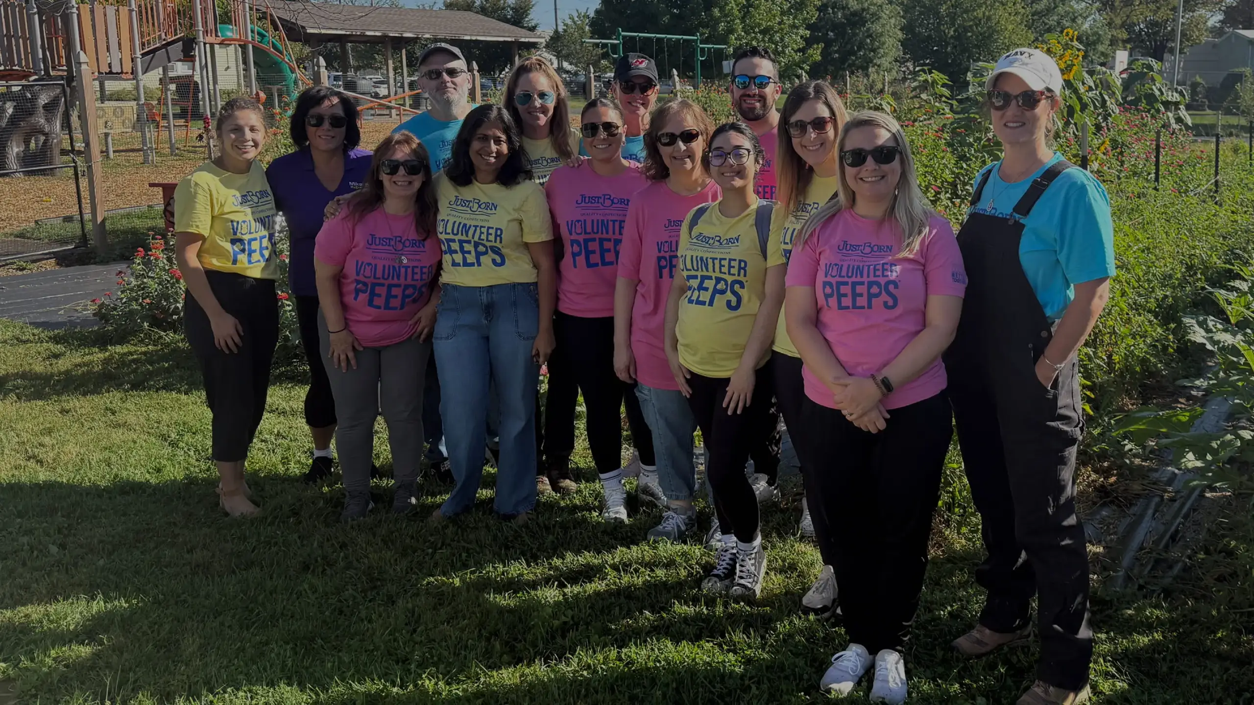 A diverse group of about 15 volunteers from Just Born Quality Confections stand together in a sunny outdoor garden. Most are wearing pink and yellow t-shirts that say "Volunteer PEEPS." They are smiling for a group photo, with a playground and sunflowers in the background.
