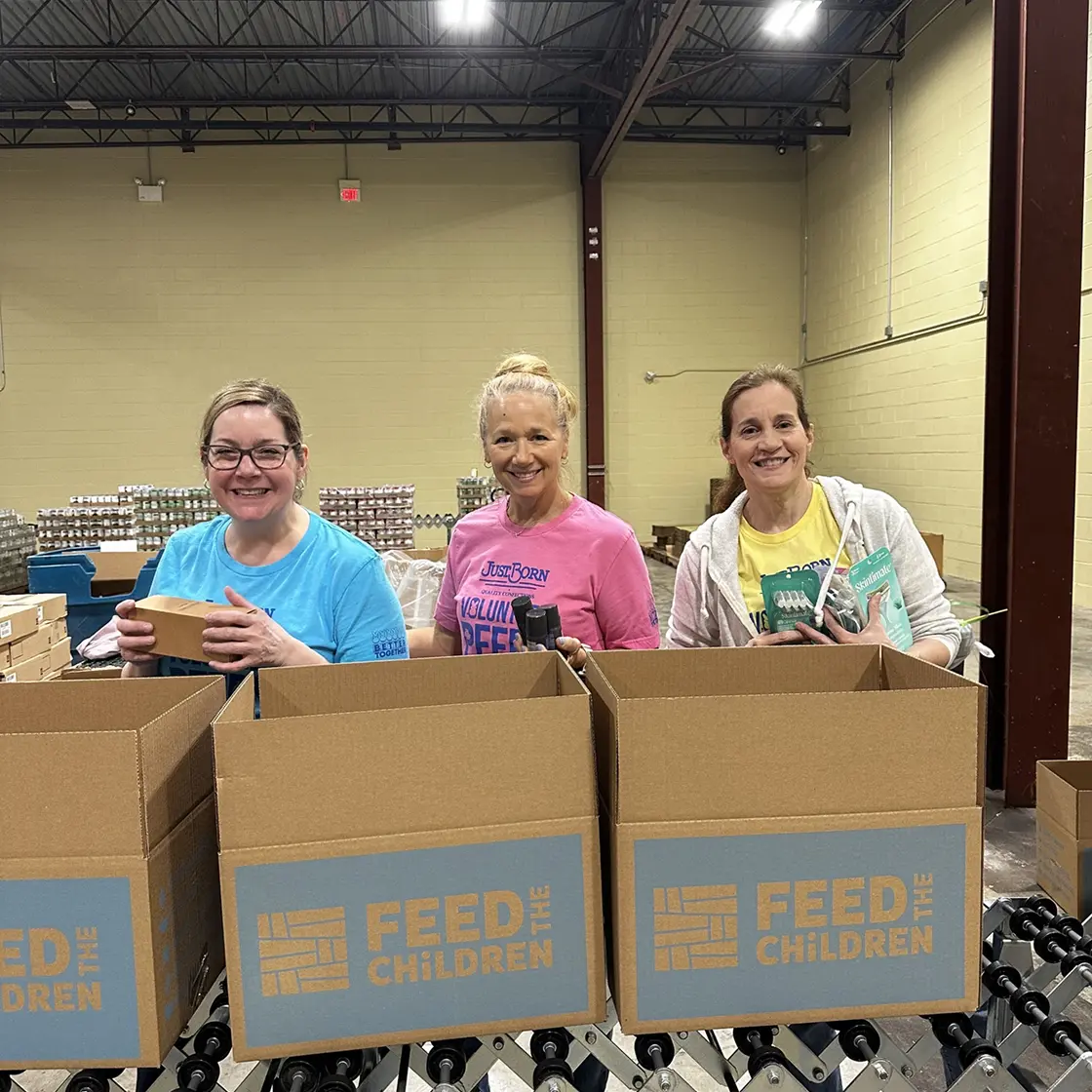 Three volunteers wearing colorful shirts stand behind large cardboard boxes labeled ‘Feed the Children’ while packing items inside a warehouse setting