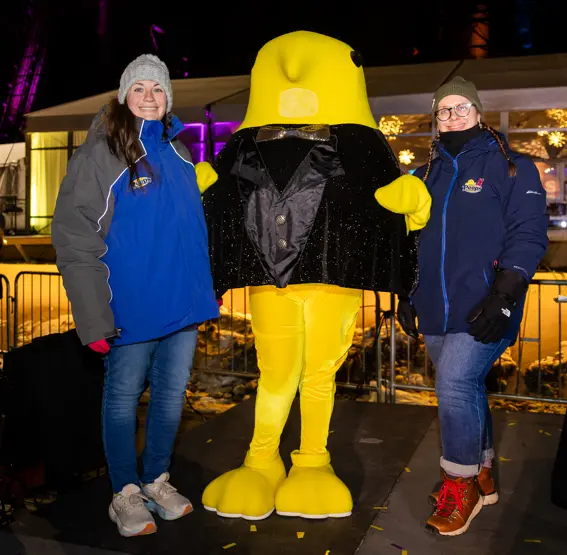 Two people pose with a yellow bird mascot at a nighttime outdoor event.